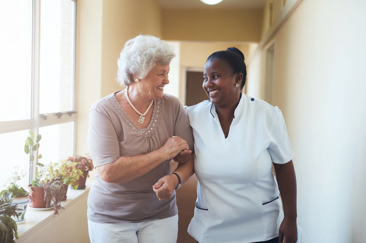 Elderly woman and caregiver sharing a cheerful moment while walking indoors.